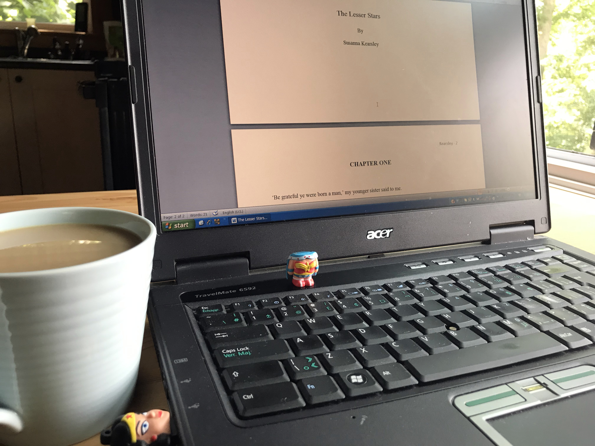 Susanna's laptop is open on a table with a cup of coffee beside it. On the screen is an open Word document with the title of the book she is beginning to write, The Lesser Stars, and the first line, "Be grateful ye were born a man, my younger sister said to me."