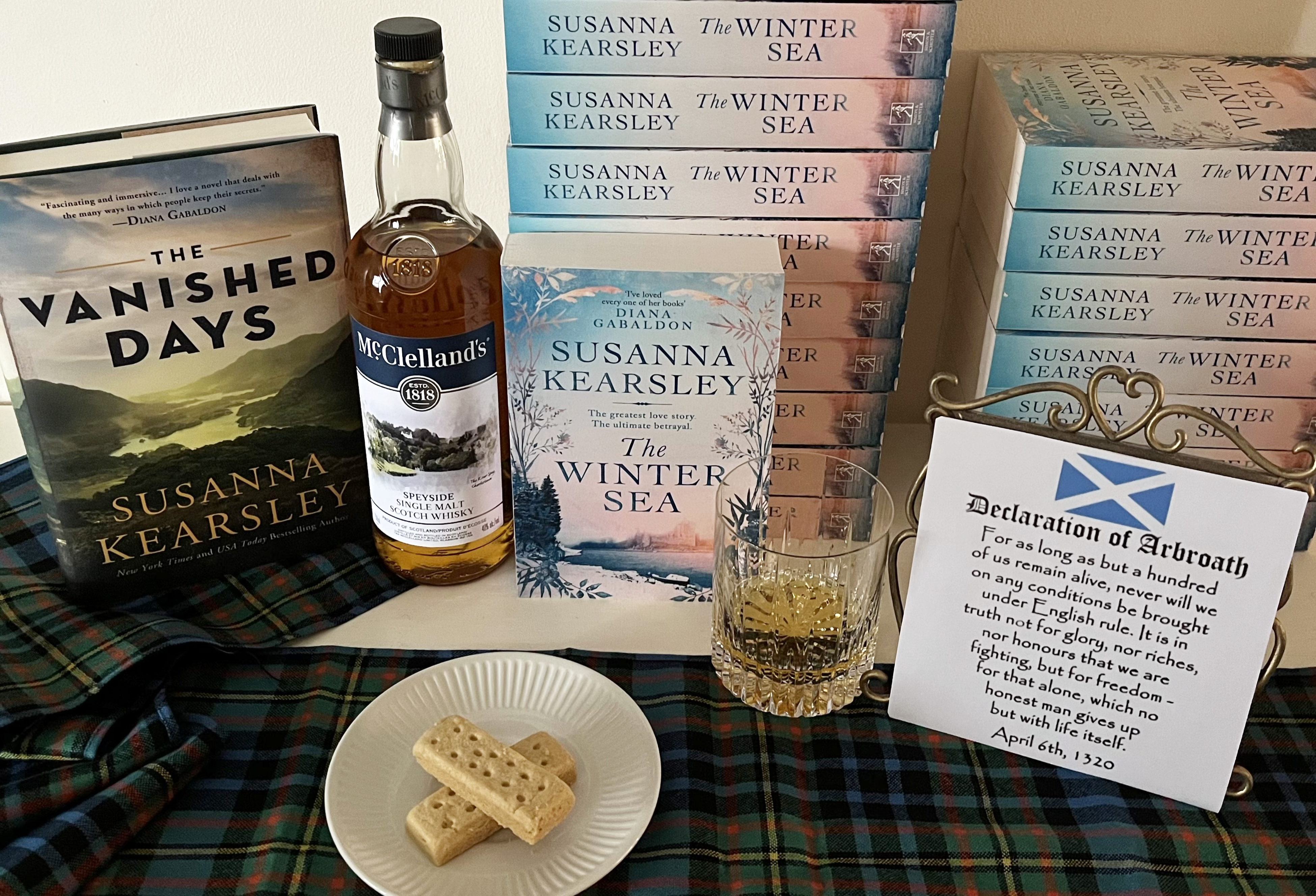 Susanna's books The Vanished Days and The Winter Sea in a still-life arrangement with a bottle and glass of whisky, shortbread biscuits crossed in a Saltire, and a copy of part of the Declaration of Arbroath, all on top of a tartan wool scarf.