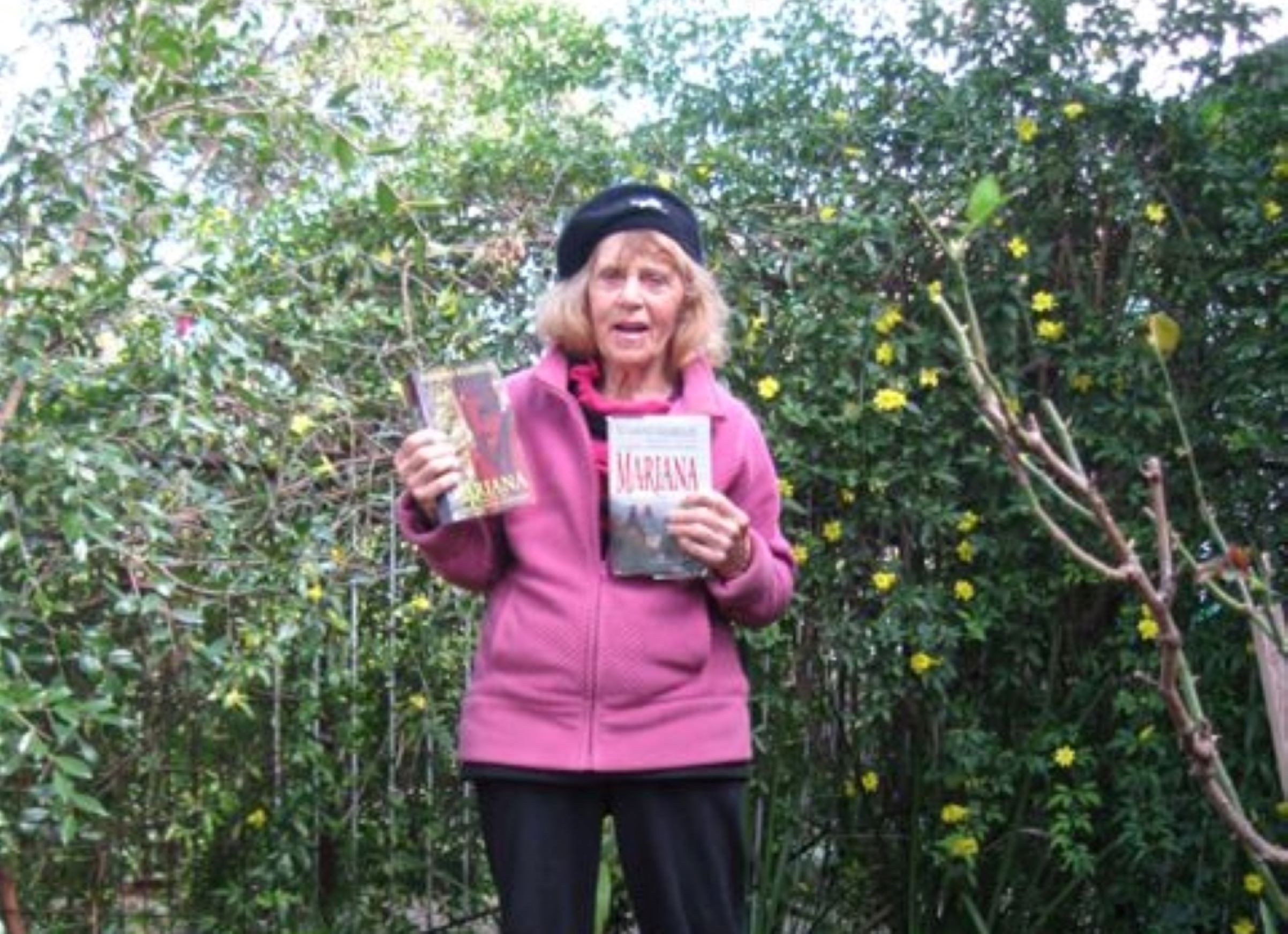 Thelma Daw stands in her garden in Australia holding two copies of Susanna Kearlsey's book Mariana. She is wearing a pink shirt and a jaunty cap.
