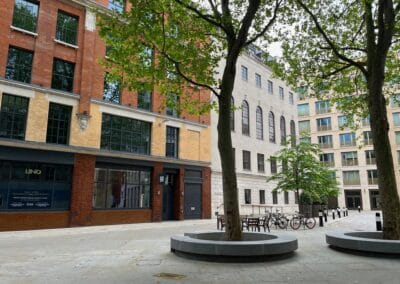 A view looking north in Bartholomew's Close, London, in spring 2023 - a deserted courtyard surrounded by tall, modern buildings, with a few tall trees remaining to cast shade over the pavement, and a small brass plaque on the pavement in the foreground.