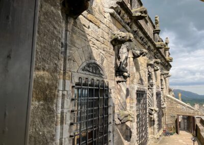 A view along the Prince's Walk at Stirling Castle, Scotland, with a variety of gargoyle-like carvings protruding from the grey stone wall that's bathed in sun, beneath a grey sky.