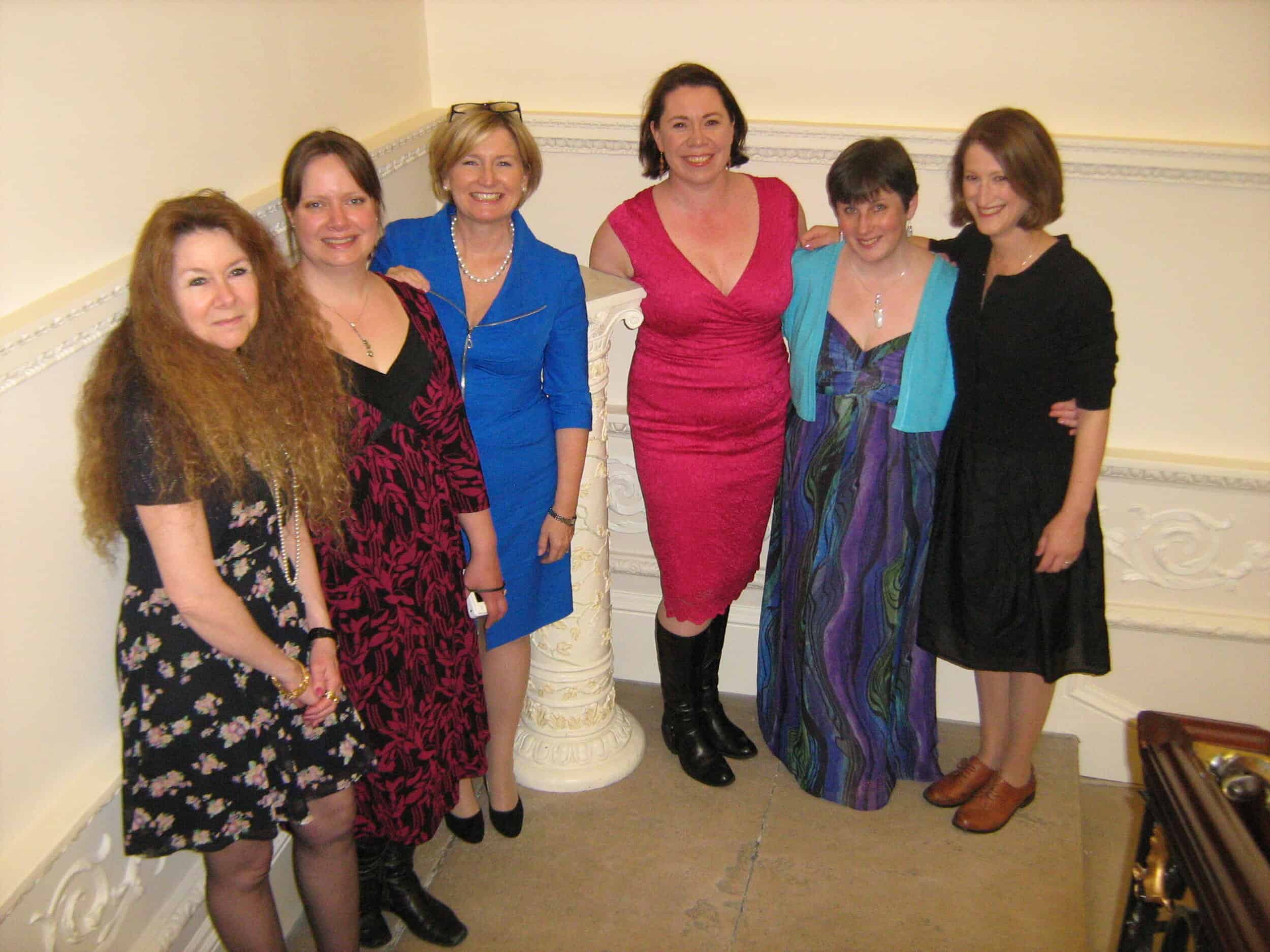 A photograph of the six members of The Heroine Addicts (former group blog), taken in 2013 in London at a Romantic Novelists' Association event. They are wearing smart semi-formal dresses and standing in a line with their arms around one another, smiling at the camera. Left to right, the writers pictured are: Christina Courtenay, Anna Louise Lucia, Liz Fenwick, Brigid Coady, Susanna Kearsley, and Julie Cohen.